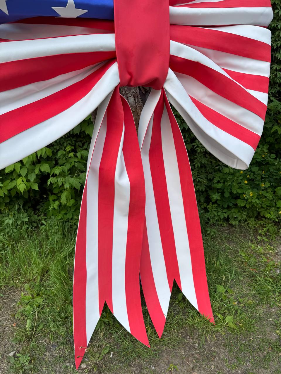 May include: A large decorative bow featuring the American flag design. The bow has red and white striped ribbons and a blue section with white stars. The bow is displayed outdoors against a backdrop of green foliage and grass.