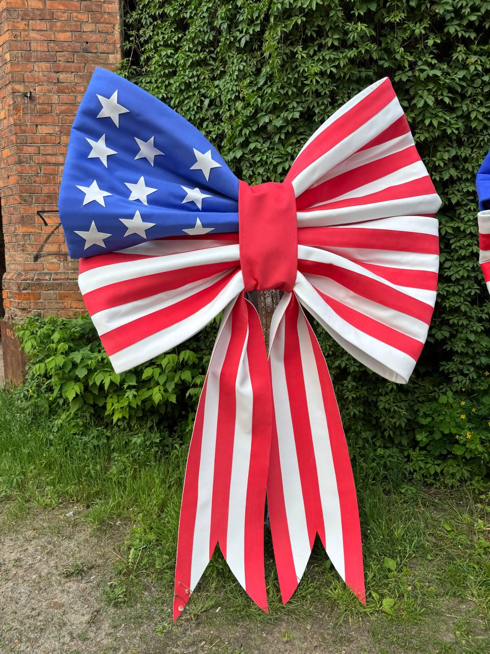 May include: Large decorative bow featuring the American flag design. The bow has a blue section with white stars, red and white stripes, and long red and white striped streamers. Perfect for patriotic decorations.