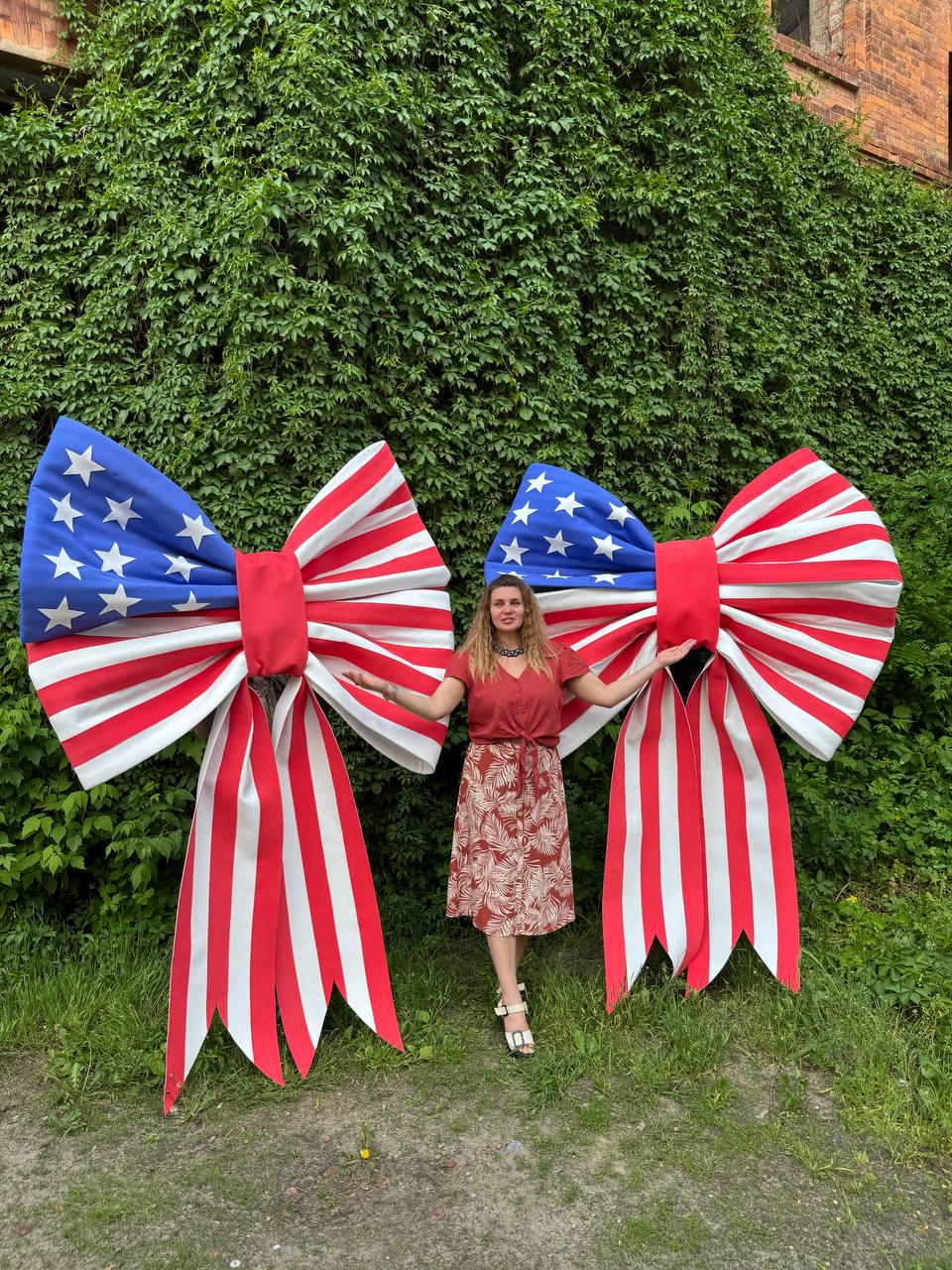 May include: Two large decorative bows in the style of the American flag. The bows feature a blue section with white stars and red and white stripes. The bows are red in the center and have long red and white striped tails. A person is standing between the bows.
