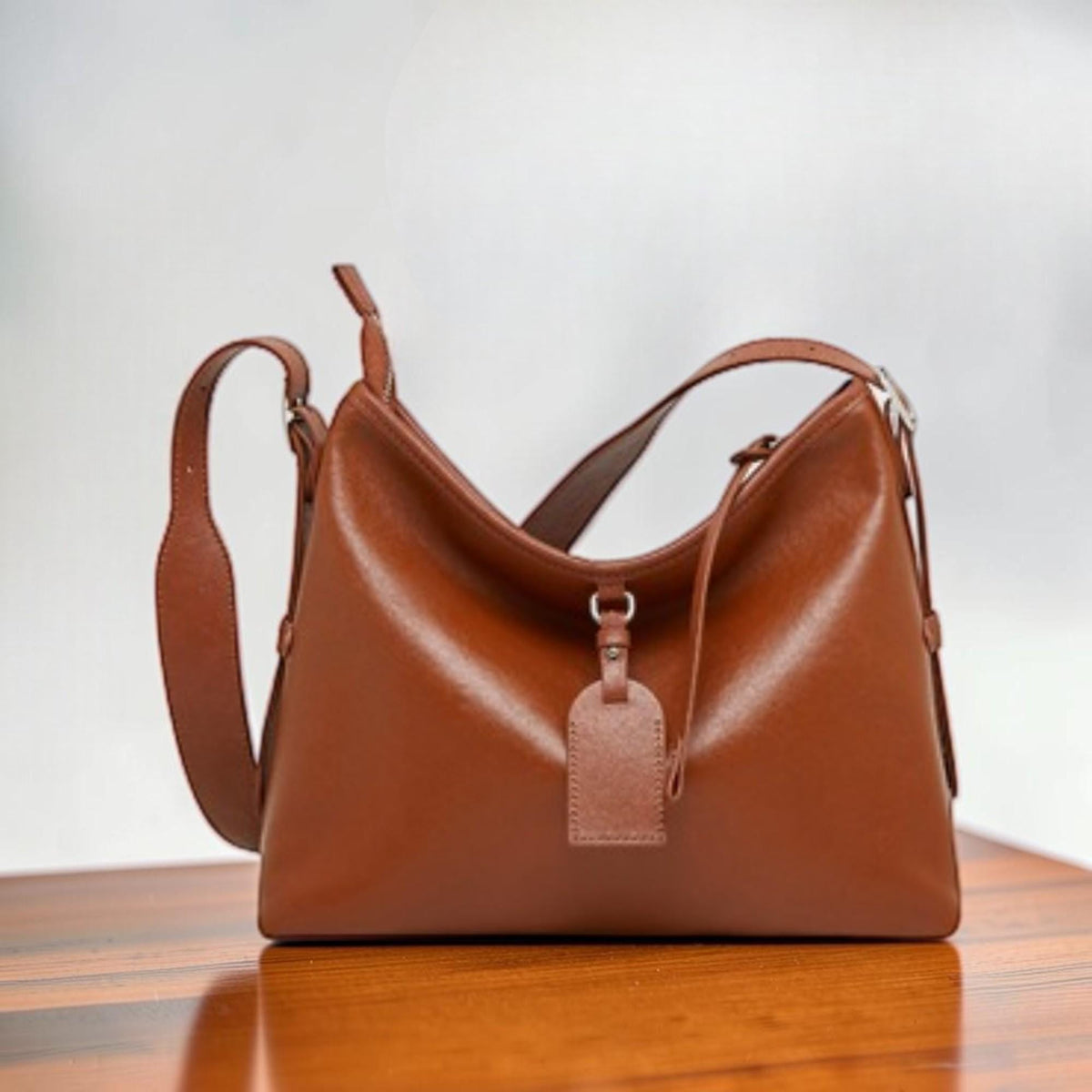 May include: A brown leather shoulder bag with a long strap and a small tag hanging from the front. The bag has a curved shape and a shiny finish. The bag is sitting on a wooden surface.