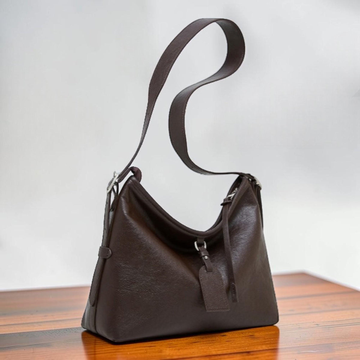May include: A dark brown leather shoulder bag with a long strap. The bag has a curved shape and a small tag hanging from the front. The bag is sitting on a wooden surface.