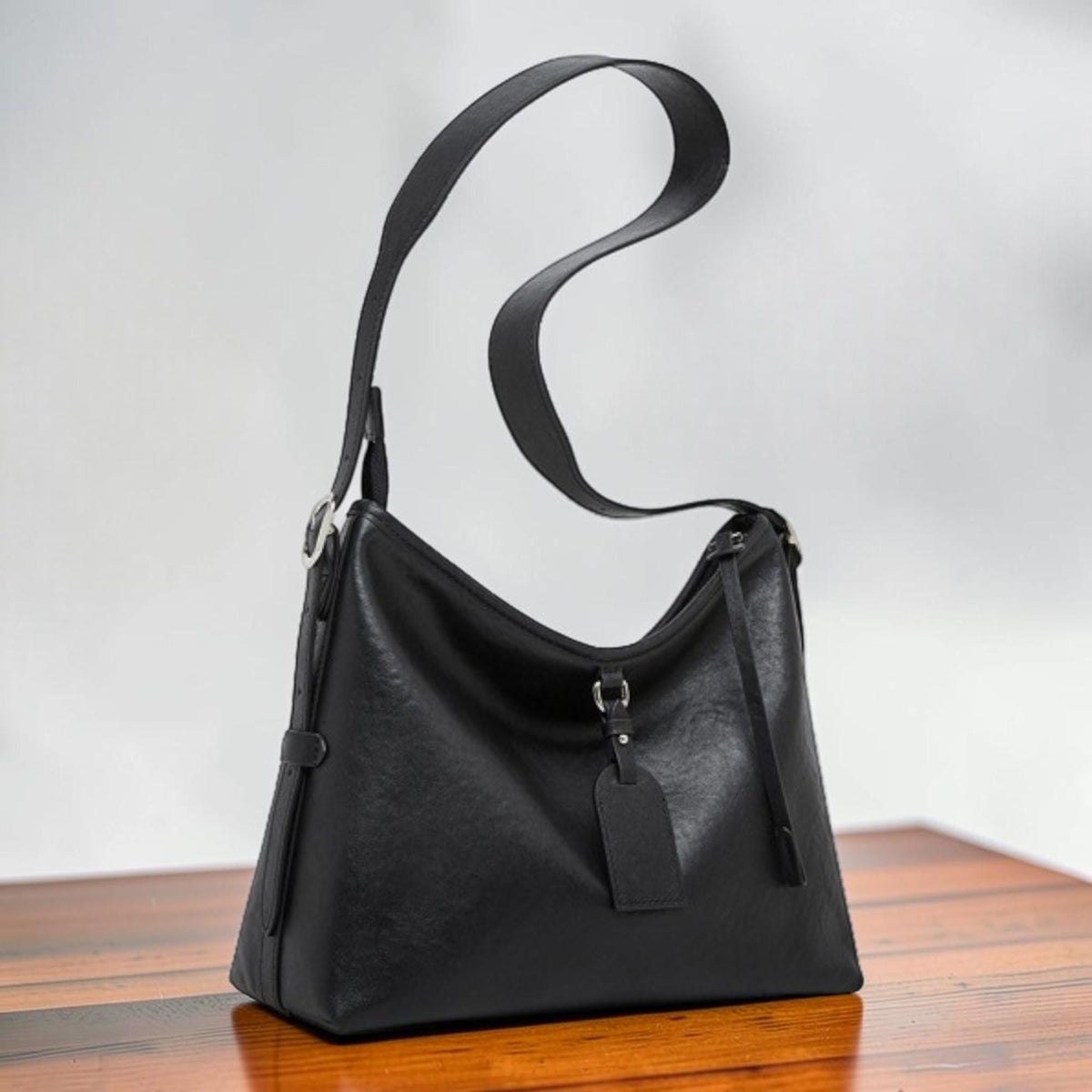 May include: A black leather shoulder bag with a long, adjustable strap. The bag has a rectangular shape with a rounded top and a small tag hanging from the front. The bag is made of a textured leather material.