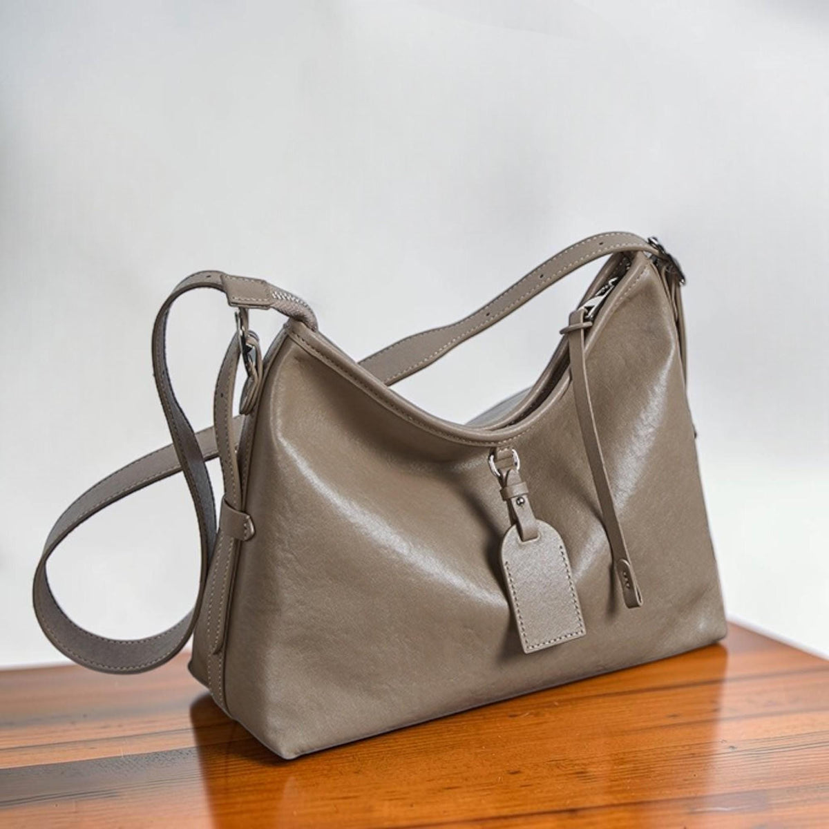 May include: A taupe-colored shoulder bag with a long, adjustable strap and a smaller handle. The bag has a rectangular tag hanging from a metal ring. The bag is made of a smooth, leather-like material and sits on a wooden surface.