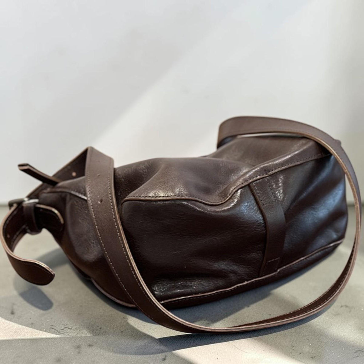 May include: A dark brown leather shoulder bag with a long strap. The bag has a rounded shape with visible stitching and a flap closure. The strap is adjustable and appears to be made of the same leather material.