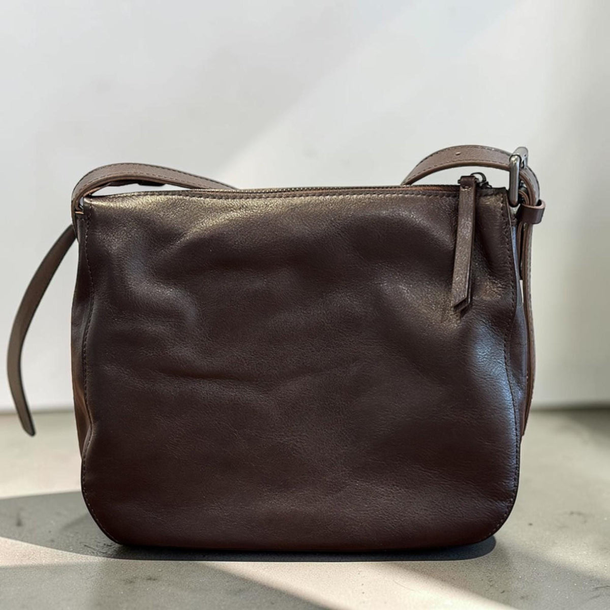 May include: A dark brown leather crossbody bag with a zipper closure and adjustable strap. The bag has a simple, classic design with a smooth texture. The strap is attached with a silver-colored buckle.