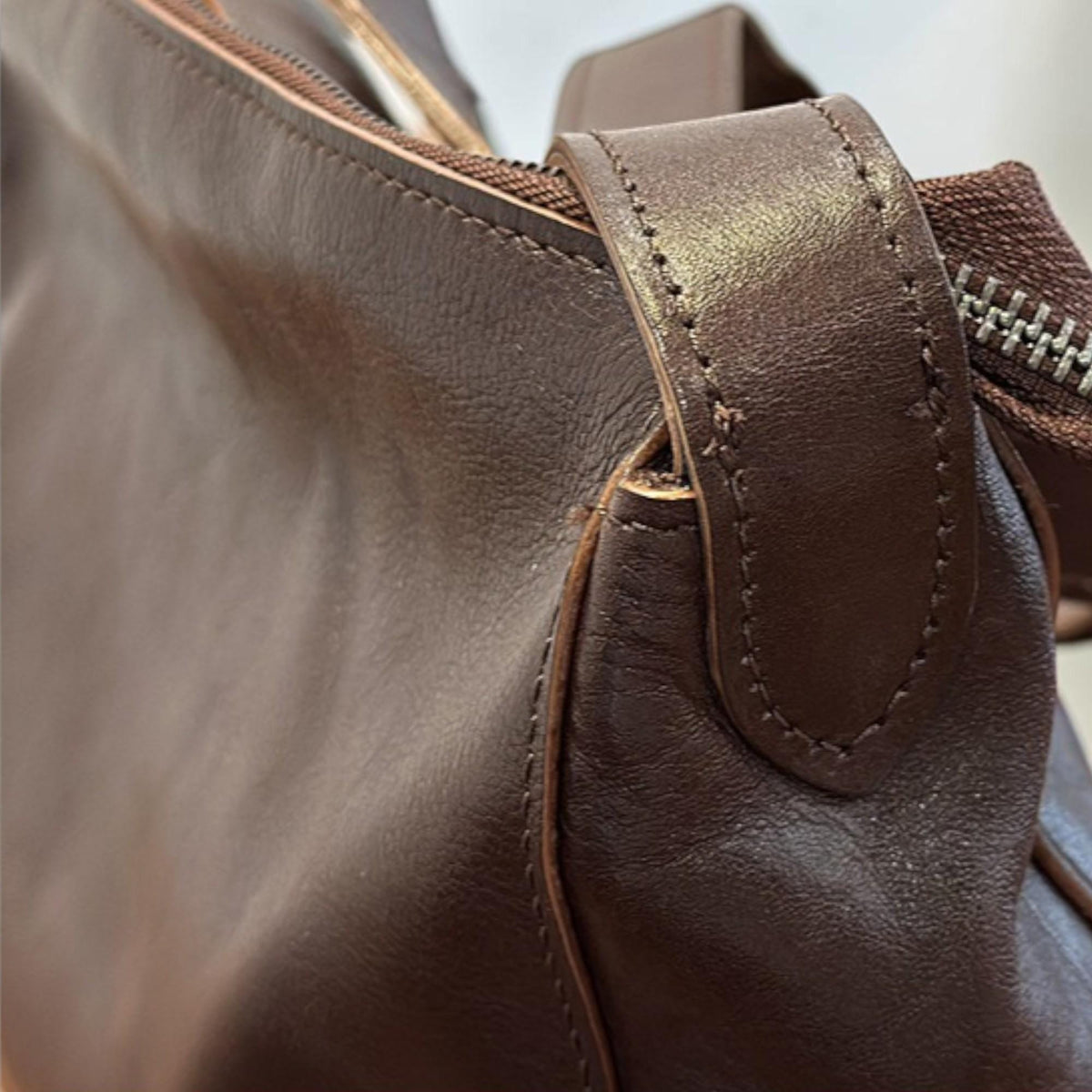 May include: Close-up of a brown leather handbag. The bag features a zipper closure and a handle. The leather appears to be smooth and has a rich, dark brown color. The stitching is visible along the seams and handle.