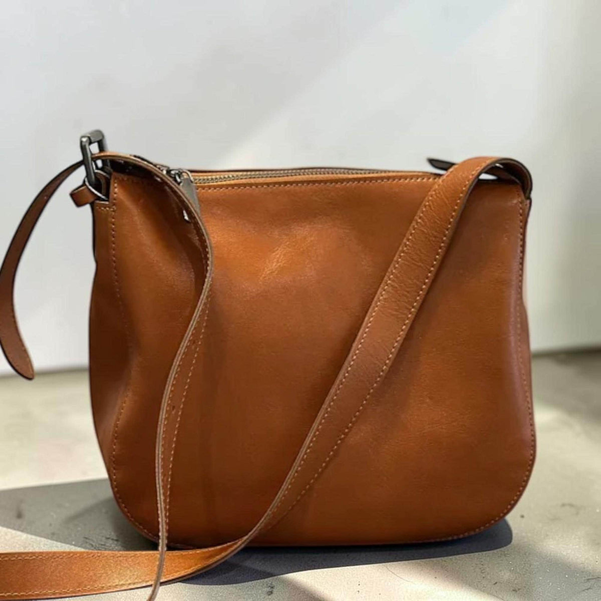 May include: A brown leather crossbody bag with a long adjustable strap. The bag has a curved bottom and a zipper closure. The leather has a smooth texture and a warm, natural color. The bag is designed for everyday use.