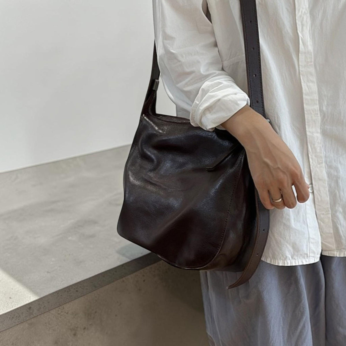 May include: A dark brown leather shoulder bag with a long adjustable strap. The bag has a soft, slouchy design and is worn over the shoulder. The bag is made of a smooth, supple leather.