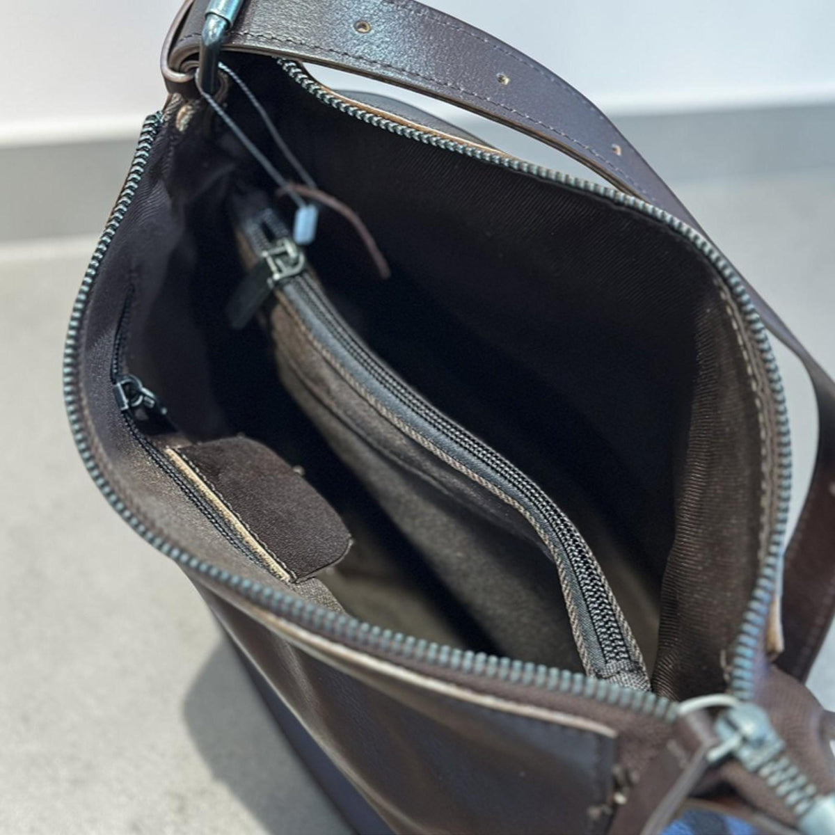 May include: A brown leather shoulder bag with a long strap. The bag has a zippered main compartment and an interior pocket. The interior is a darker brown. The bag is open, revealing the inside.