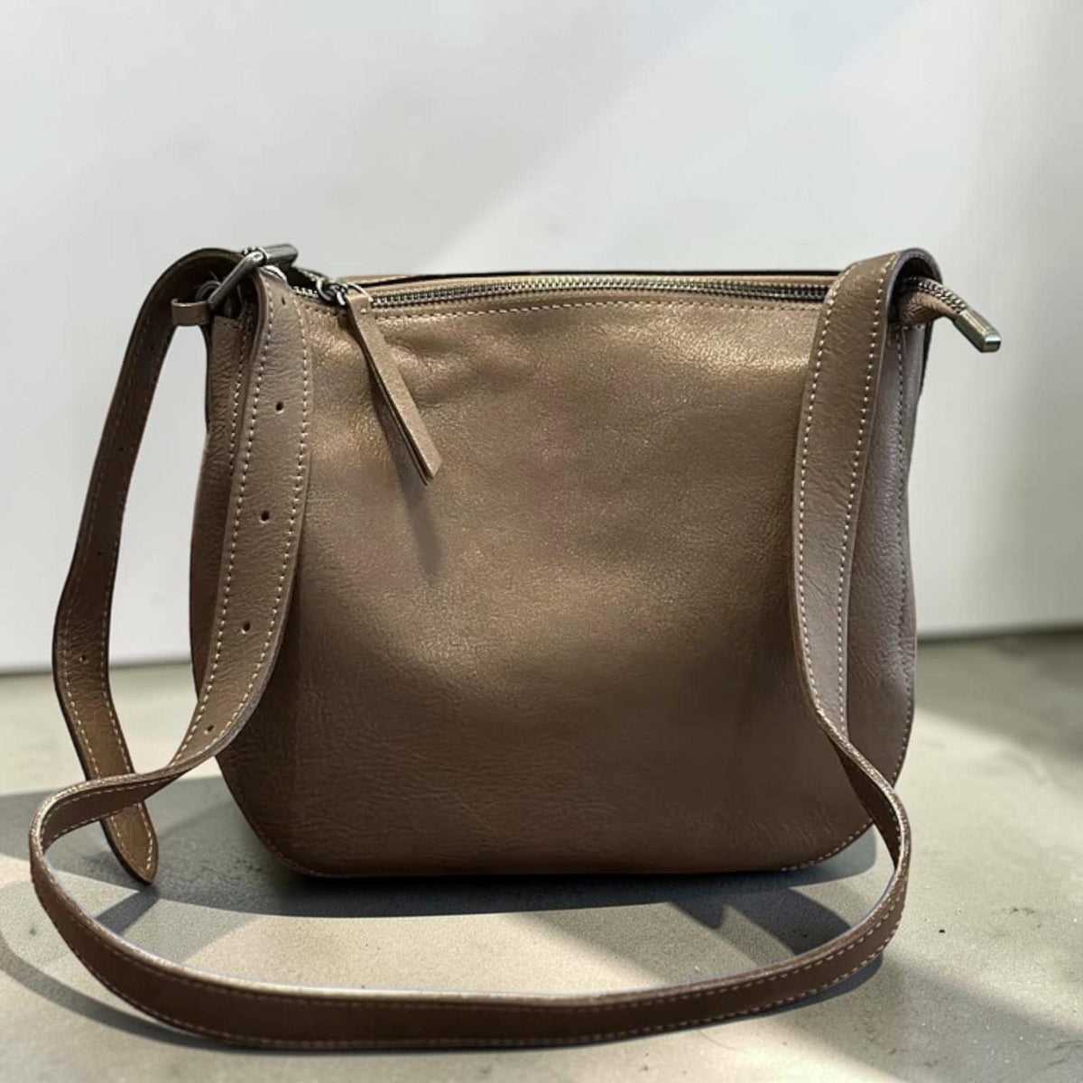 May include: A taupe-colored leather crossbody bag with a zipper closure and adjustable strap. The bag has a rounded shape and is made of a soft, textured leather. The strap is attached with silver-toned hardware.