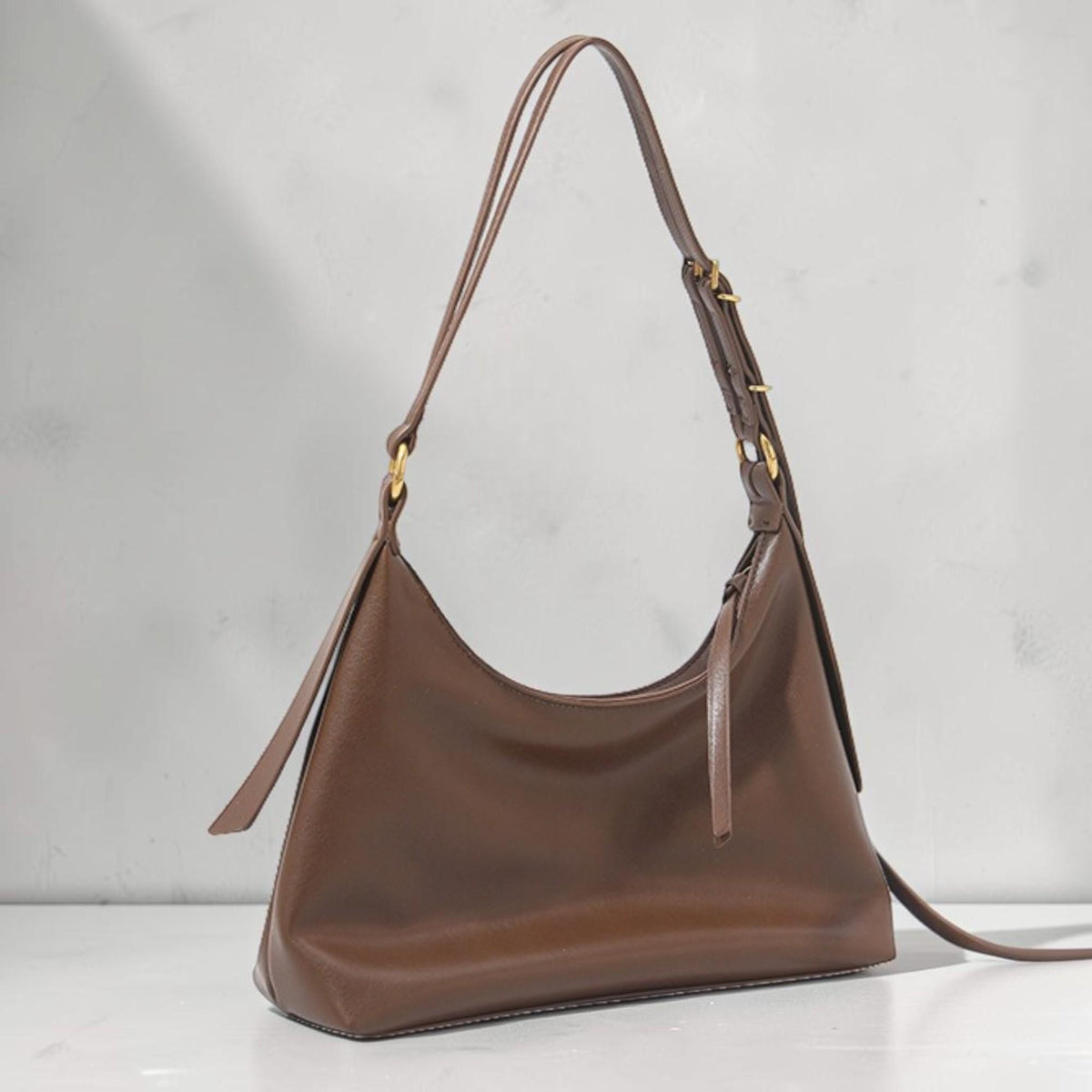 May include: A brown leather shoulder bag with a curved shape. The bag has a long strap and gold-colored hardware. The bag is made of a smooth, matte leather and is suitable for everyday use.
