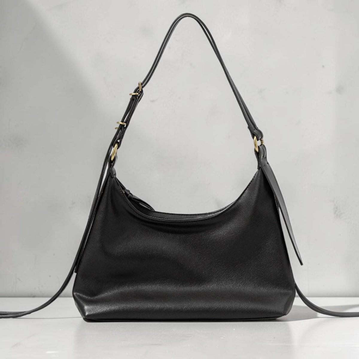 May include: A black shoulder bag with a curved design. The bag has a long, adjustable strap and gold-colored hardware. The bag is made of a smooth material and has a zipper closure.