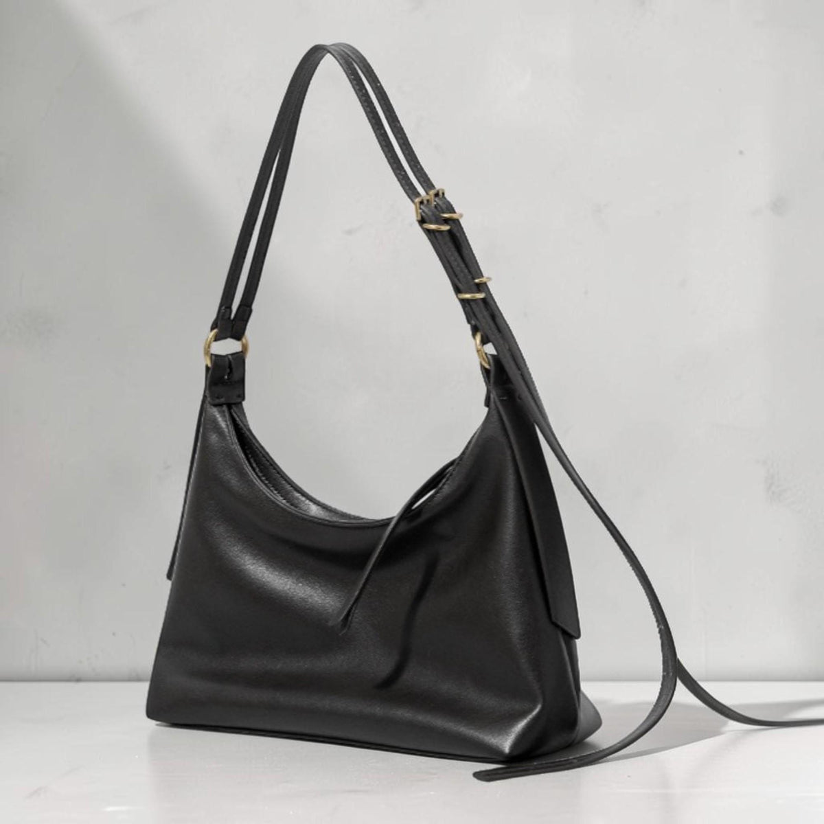 May include: A black leather shoulder bag with a curved design. The bag has a long, adjustable strap with gold-colored metal hardware. The bag is shown against a neutral background.