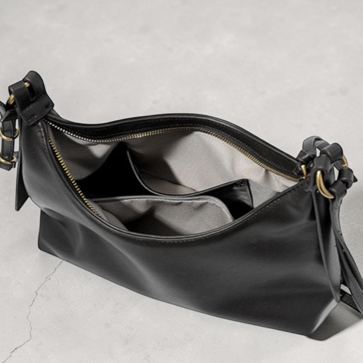 May include: Black leather shoulder bag with a zippered top and interior pockets. The bag has gold-colored hardware and a woven handle detail. The interior lining is a light gray fabric. The bag is open, revealing the inside.