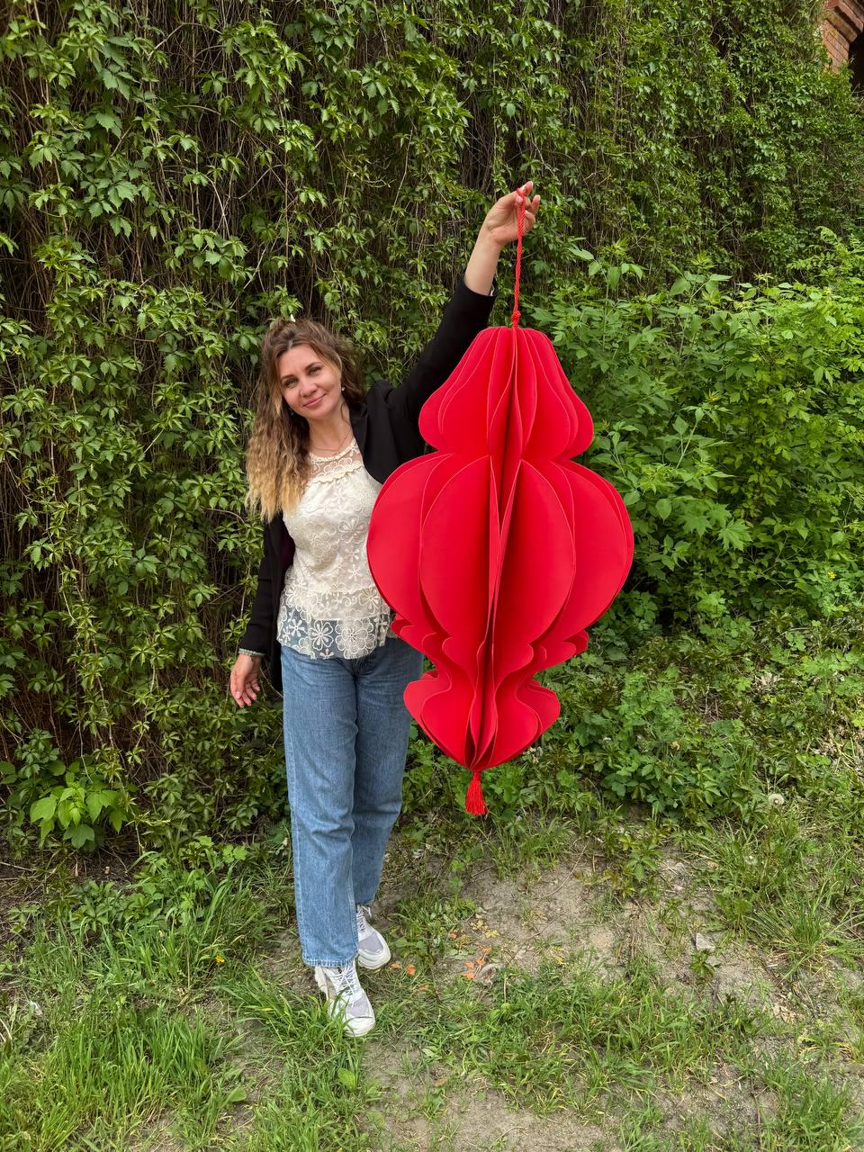 May include: A large, vibrant red paper lantern with a tassel, suspended by a red string. The lantern has a layered, three-dimensional design. The background features lush green foliage and a person.