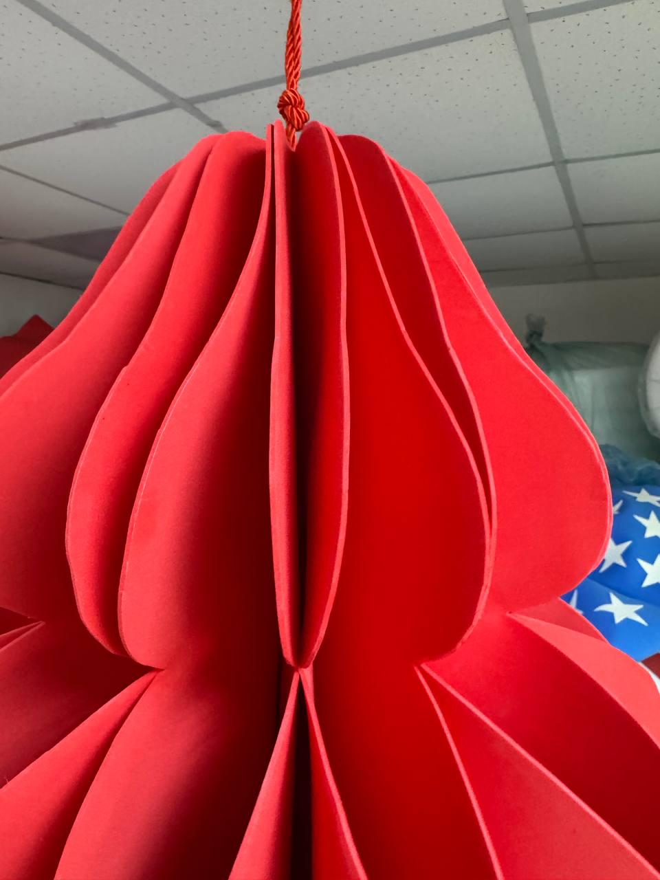 May include: A large, vibrant red paper lantern, suspended from a red cord. The lantern has a layered, petal-like design, creating a three-dimensional effect. The background shows a glimpse of a blue and white American flag.