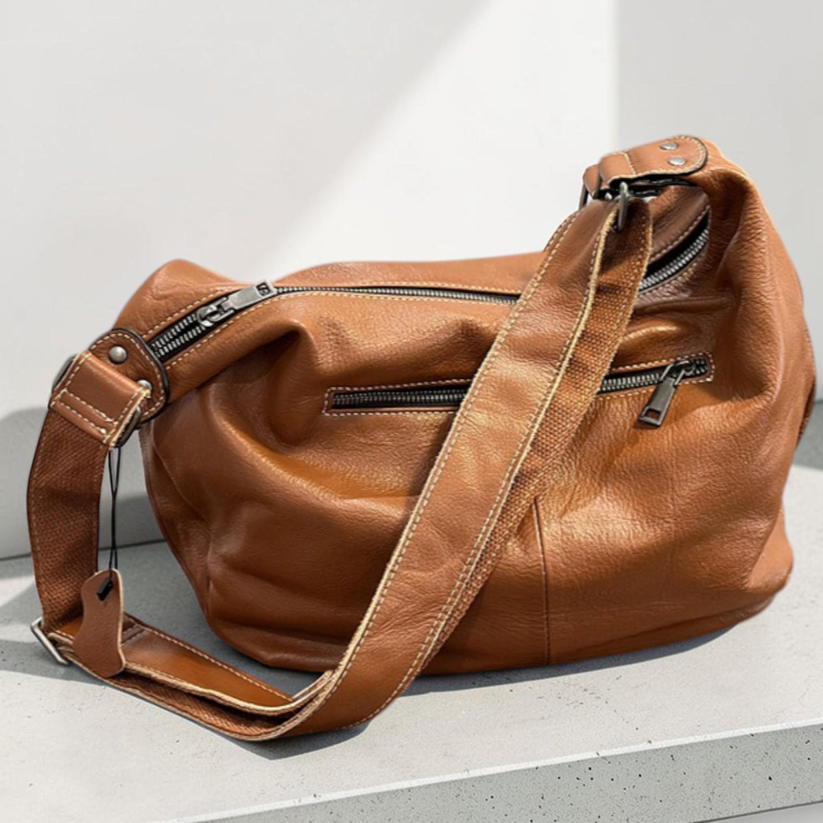 May include: A brown leather shoulder bag with a long adjustable strap. The bag has multiple zippered compartments and a textured surface. The stitching is visible in white thread. The bag is sitting on a white surface.