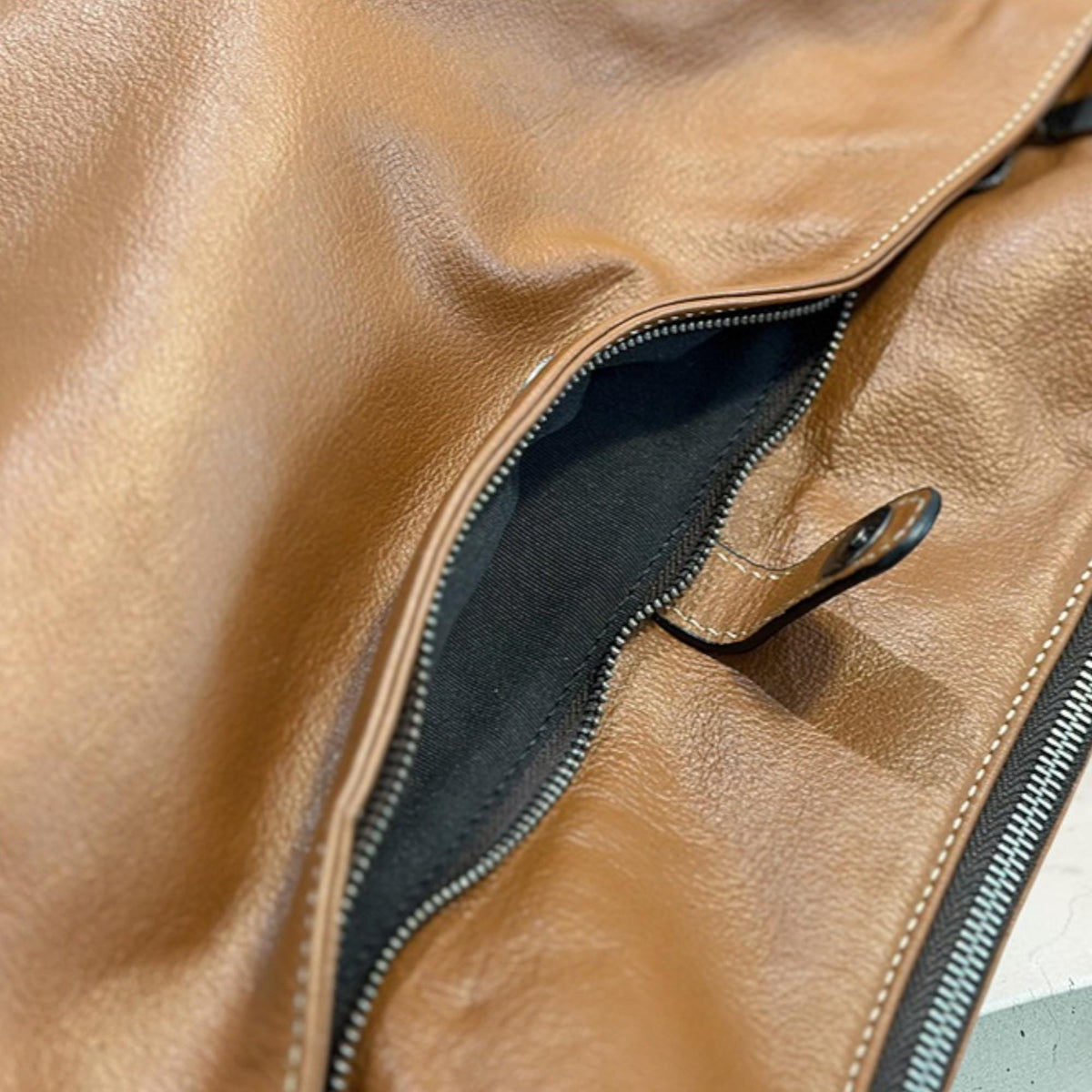 May include: Close-up of a brown leather bag with a black interior pocket. The bag features a silver zipper and a small leather tab with a metal snap closure. The leather has a smooth, slightly glossy finish.
