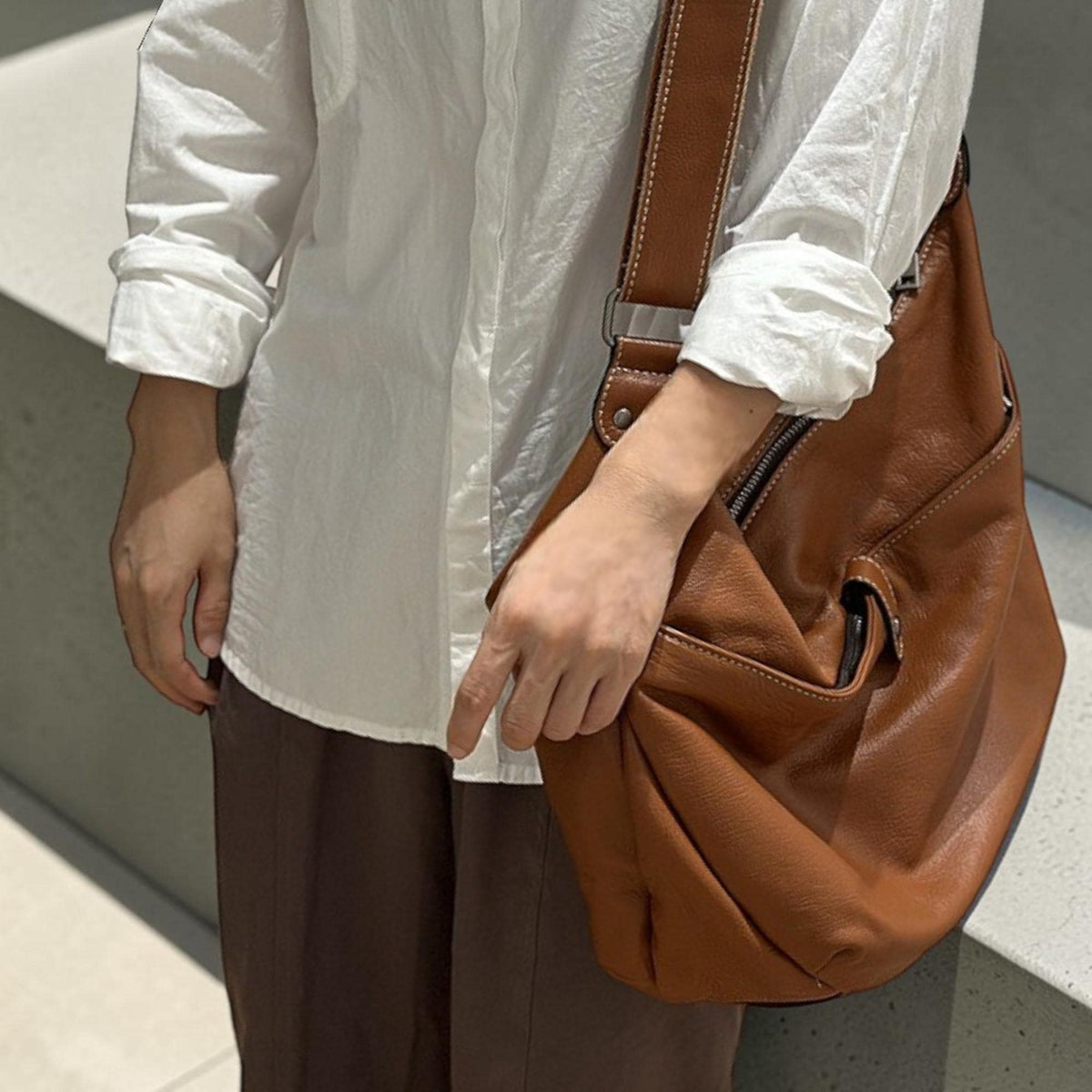 May include: A brown leather shoulder bag with a zipper closure and an adjustable strap. The bag has a pocket on the front and is worn over the shoulder. The bag is a medium size and appears to be made of soft leather.