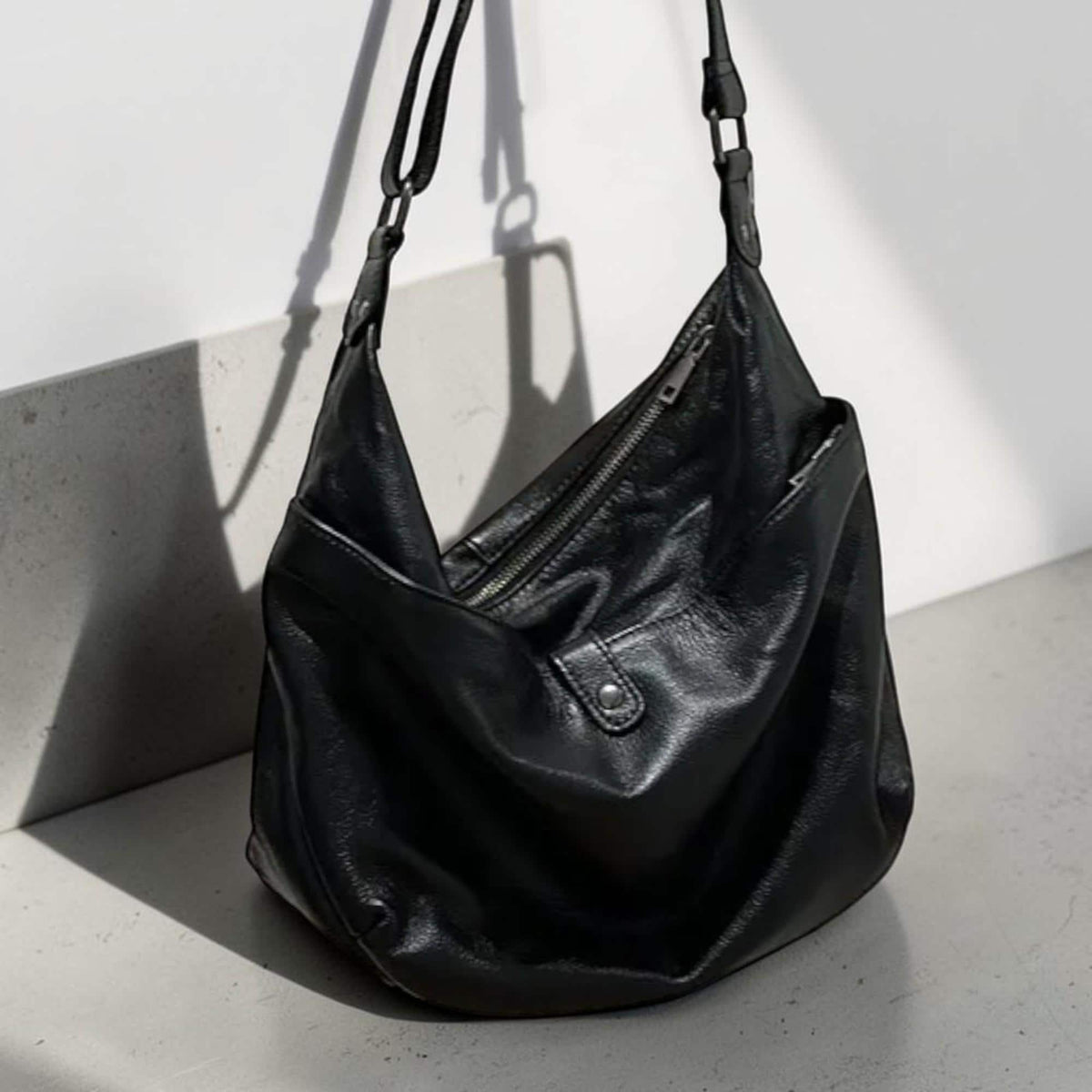 May include: Black leather shoulder bag with a slouchy design. The bag features a zippered top closure, an exterior pocket, and adjustable straps. The bag is hanging against a white wall and a gray surface.