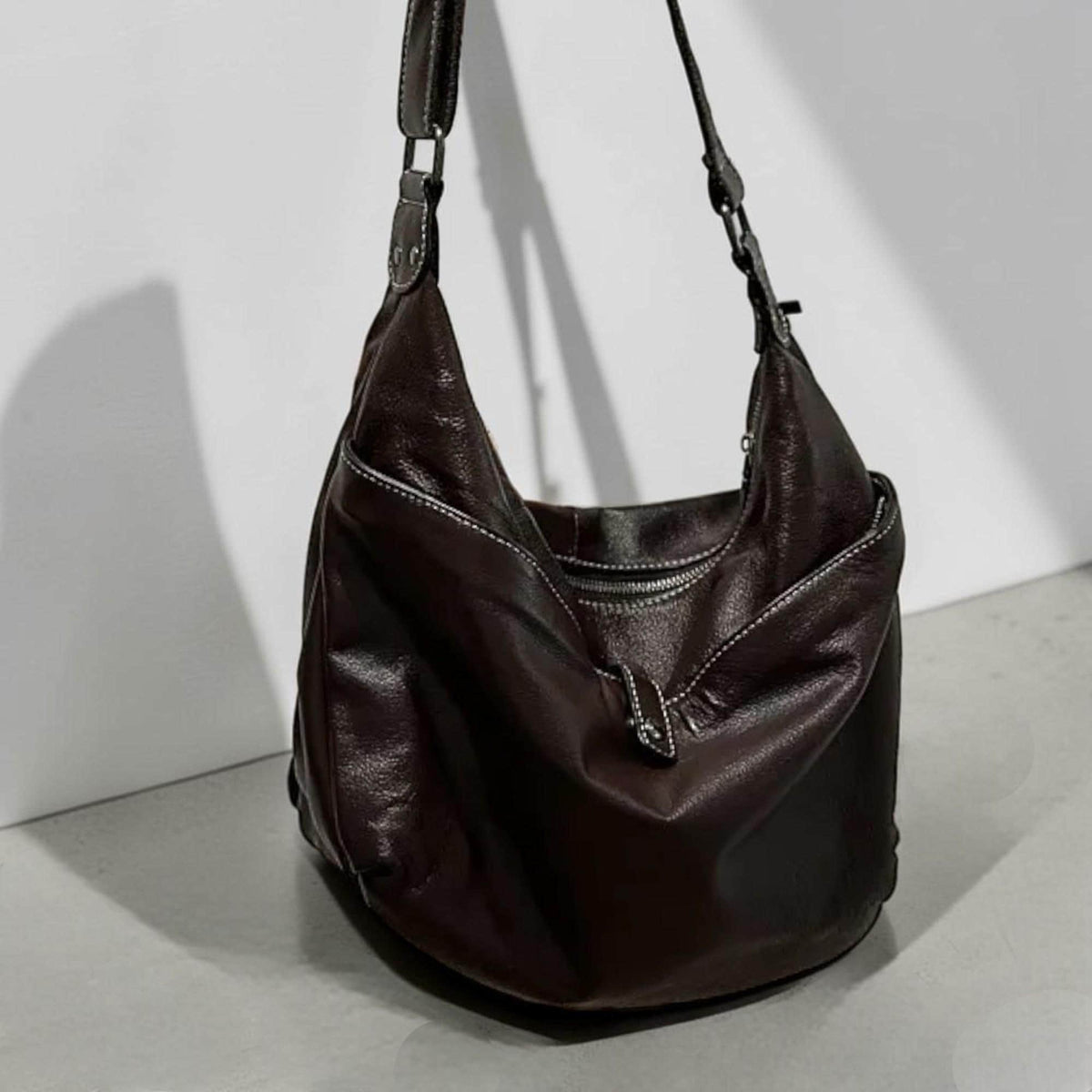 May include: A dark brown leather hobo bag with a single shoulder strap. The bag features white stitching along the seams and a zipper closure. The bag is hanging against a white wall.