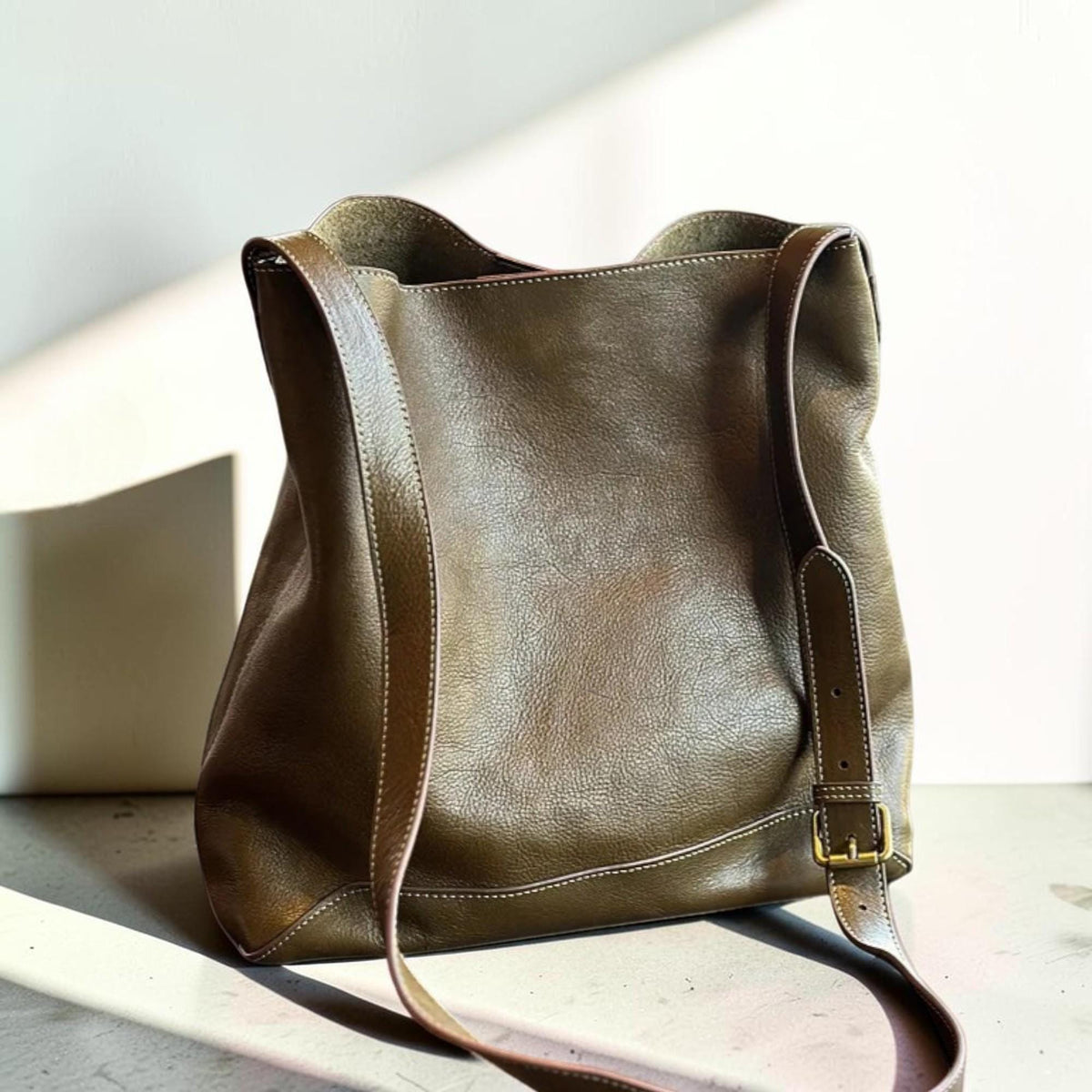 May include: Olive green leather shoulder bag with a long adjustable strap and a brass buckle. The bag has a spacious interior and is stitched with white thread. The bag is made of a soft, textured leather.