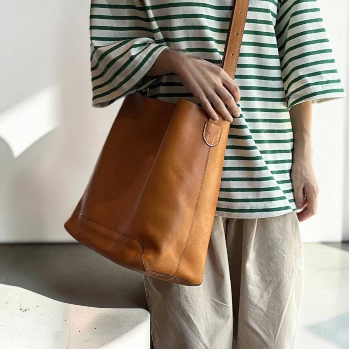 May include: A tan leather tote bag with a long shoulder strap. The bag is held by a person wearing a green and white striped shirt and tan pants. The bag has a simple, classic design.