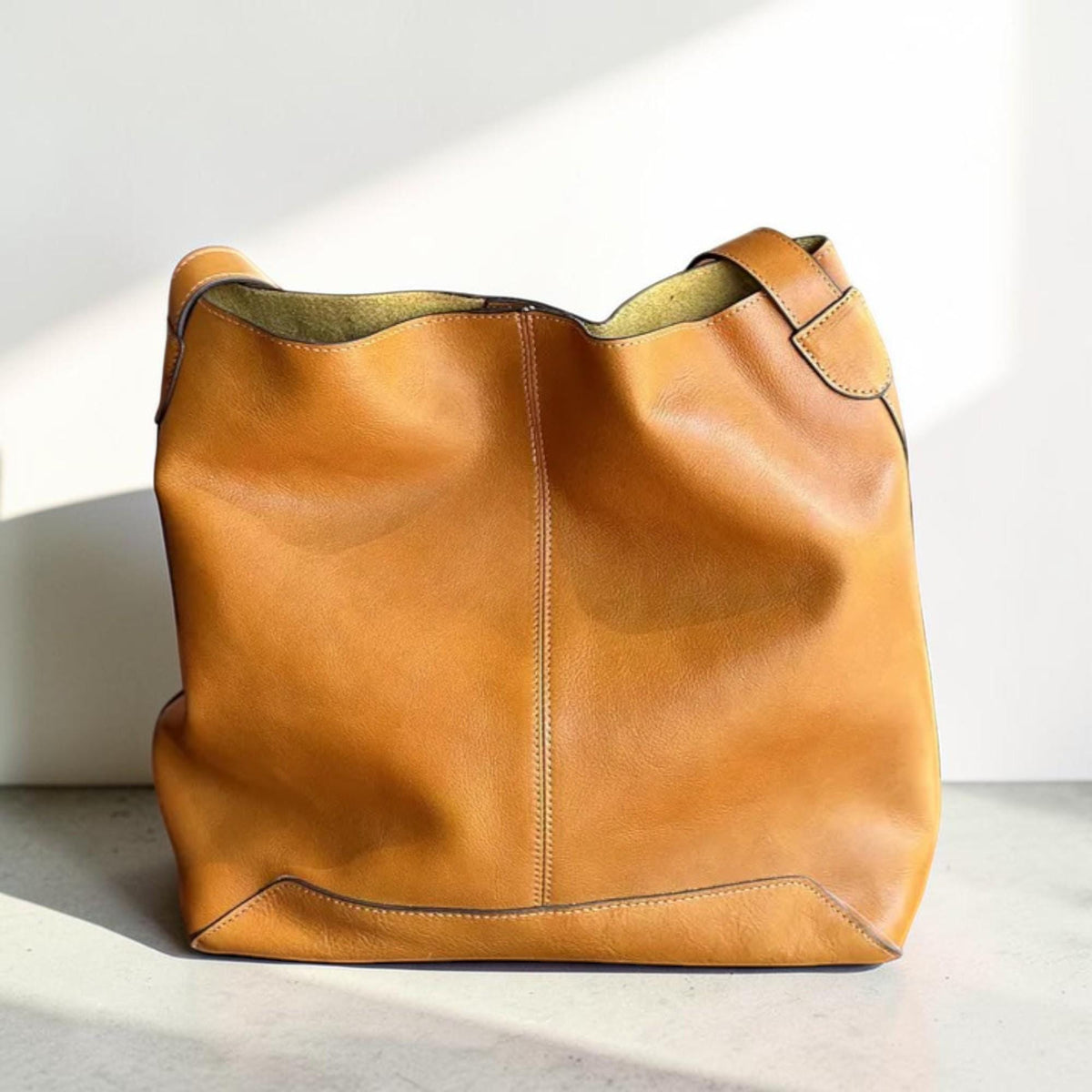 May include: A large, tan leather tote bag with a single shoulder strap. The bag has a rectangular base and a wide opening. The leather has visible grain and stitching details. The interior lining is a contrasting olive green color.