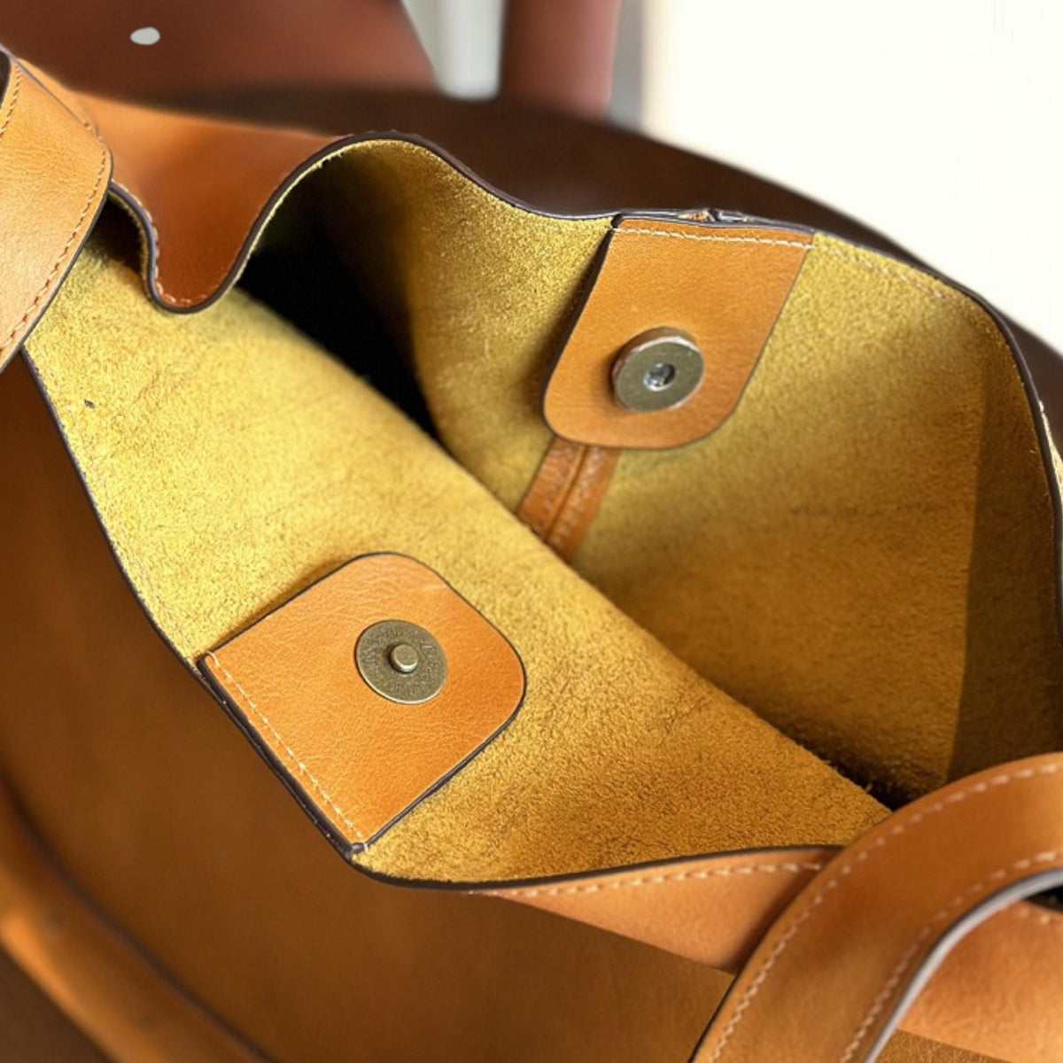 May include: Close-up of a tan leather tote bag interior. The bag features a mustard yellow suede lining and a magnetic snap closure. The bag's exterior is a lighter tan color, with a darker brown trim.