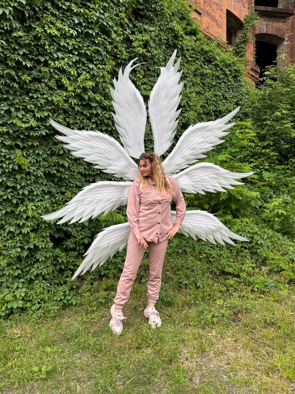 May include: A person wearing a pink outfit and white sneakers stands in front of large white angel wings. The wings are made of feathers and are arranged in a starburst pattern. The background features lush green foliage and a brick building.