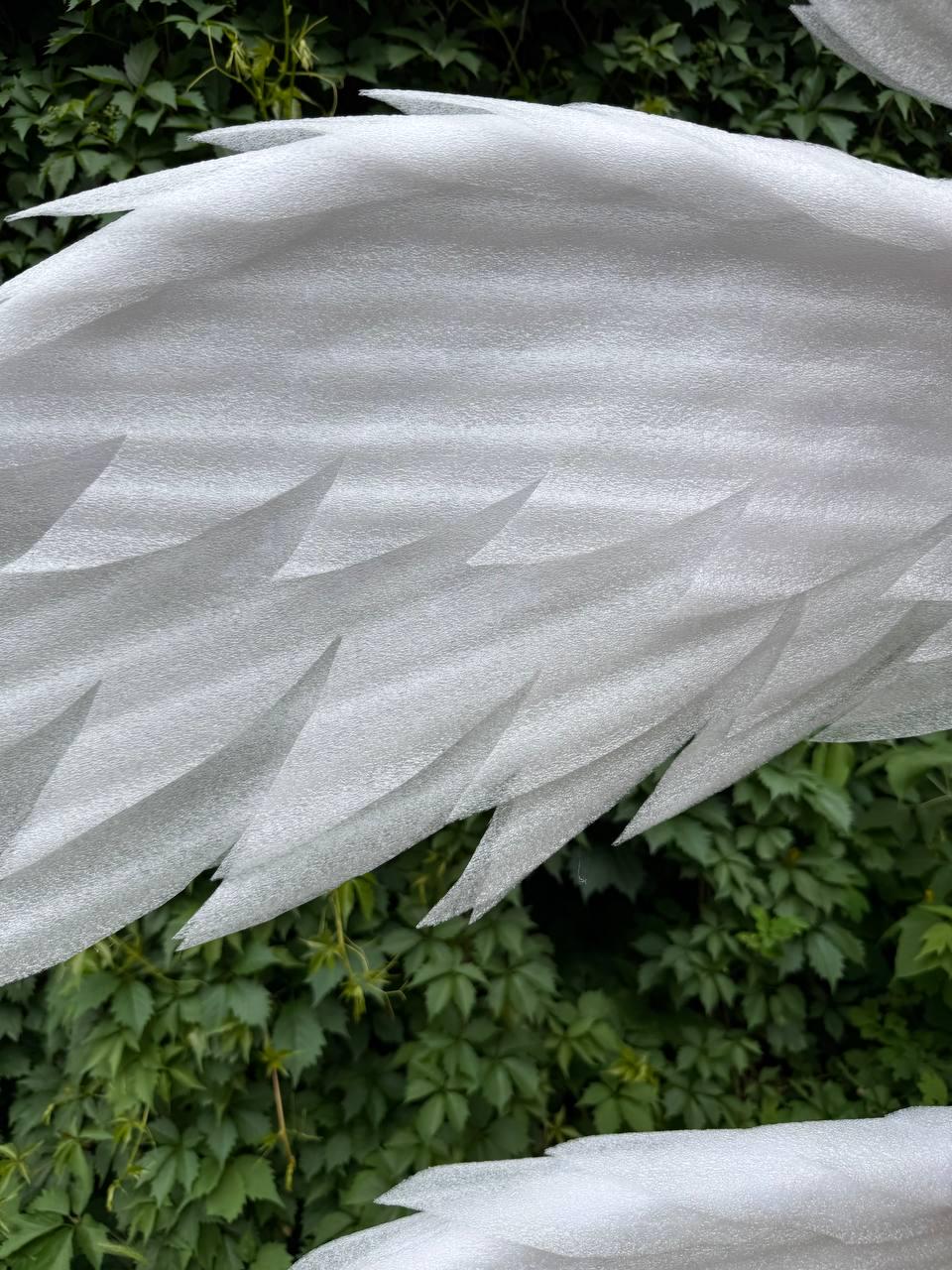 May include: Close-up of a white, textured foam object with sharp, triangular edges. The object is set against a backdrop of green foliage. The foam appears to be a protective packaging material.
