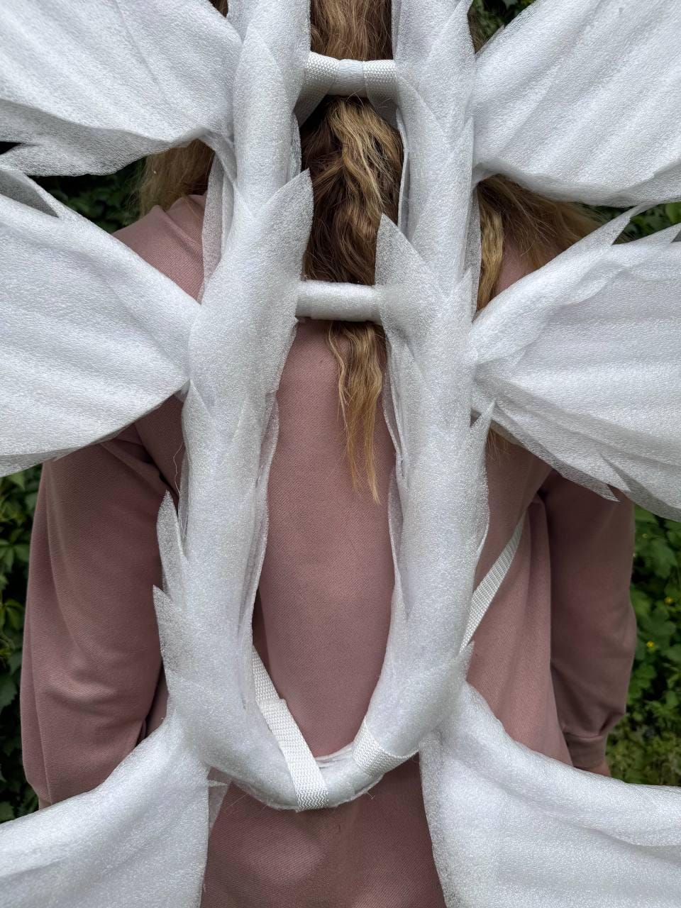 May include: A person wearing a pink sweatshirt with white packaging material wrapped around the back and shoulders. The packaging is made of a soft, white foam material and is held together with white straps.