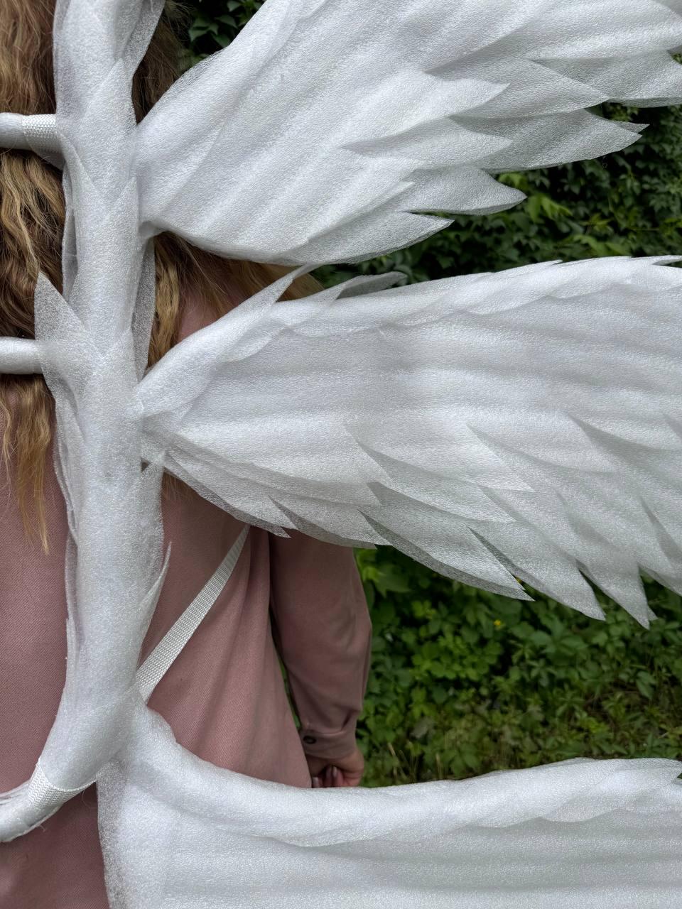 May include: White angel wings with layered feather details, wrapped in protective packaging. The wings are attached to a frame with a white strap. The background shows green foliage and a person wearing a pink shirt.