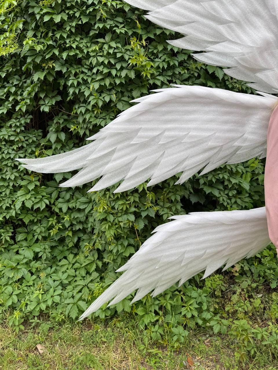 May include: Large, white angel wings with a layered feather design. The wings are made of a lightweight material and are displayed against a backdrop of green foliage and grass. The wings are a costume or decorative item.