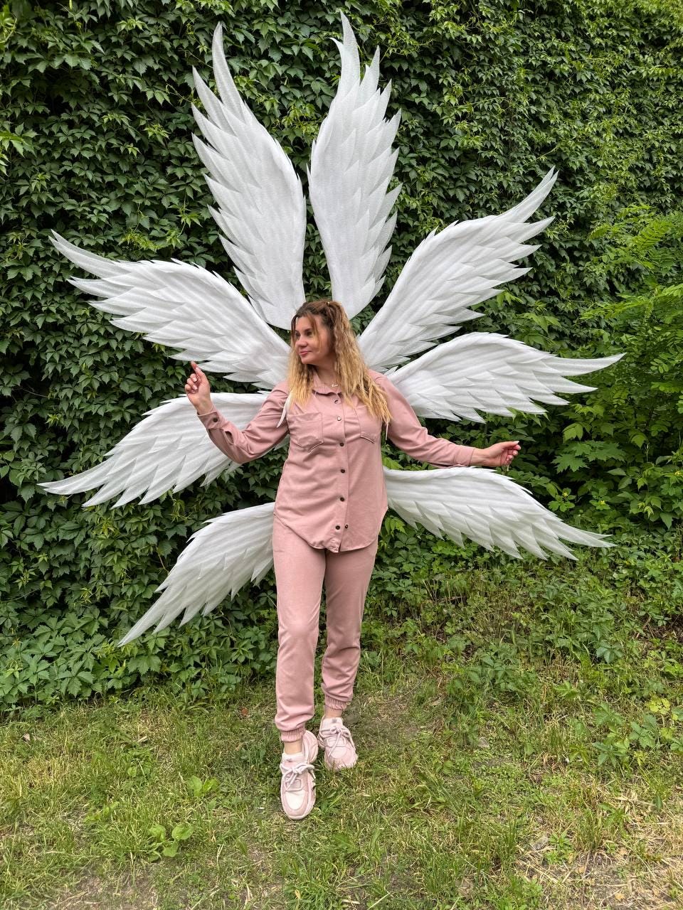 May include: A person wearing a pink outfit and white angel wings stands in front of a green leafy wall. The wings are large and detailed, with multiple layers of white feathers. The outfit includes a button-down shirt, pants, and sneakers.