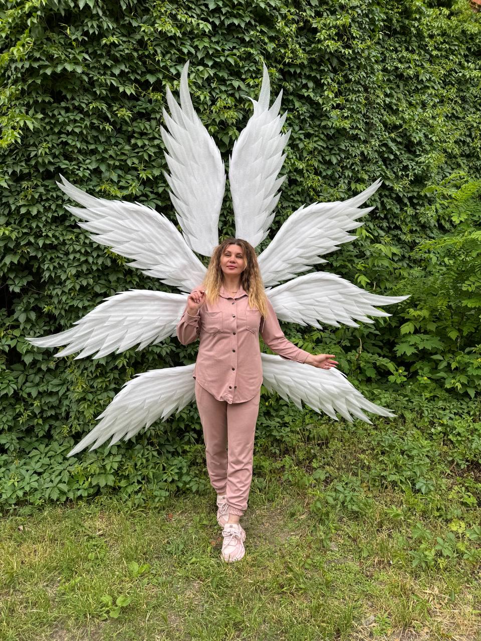 May include: A person wearing a pink outfit and white sneakers stands in front of large, white angel wings. The wings are made of layered feathers and are spread out behind the person. The background is a wall of green foliage and grass.