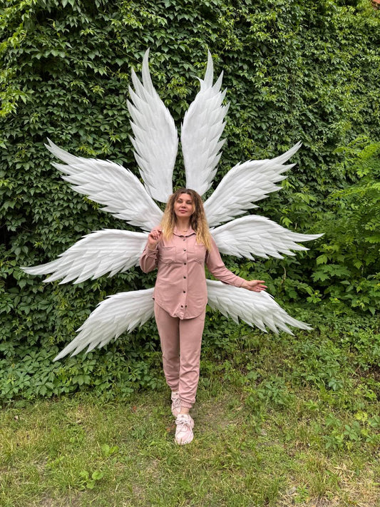 May include: A person wearing a pink outfit and white sneakers stands in front of large, white angel wings. The wings are made of layered feathers and are spread out behind the person. The background is a wall of green foliage and grass.