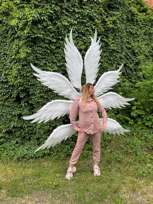 May include: A person wearing a pink outfit and white angel wings stands in front of a wall of green foliage. The wings are large and detailed, with multiple layers of white feathers. The outfit includes a button-down shirt and matching pants.