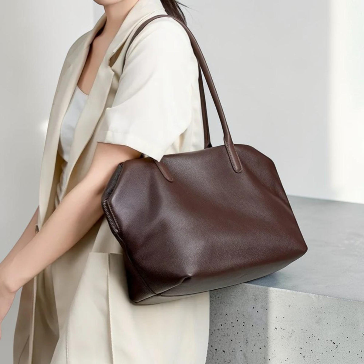 May include: A dark brown leather tote bag with long handles. The bag has a rectangular shape with a zipper closure and is carried over the shoulder. The bag is a neutral color and can be used for everyday use.