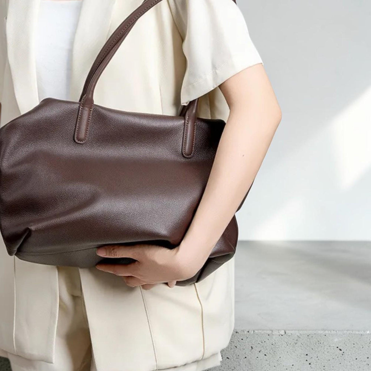 May include: A dark brown leather tote bag with two top handles. The bag has a rectangular shape with a slightly rounded top and a textured surface. The handles are a darker shade of brown and are attached to the bag with reinforced stitching.