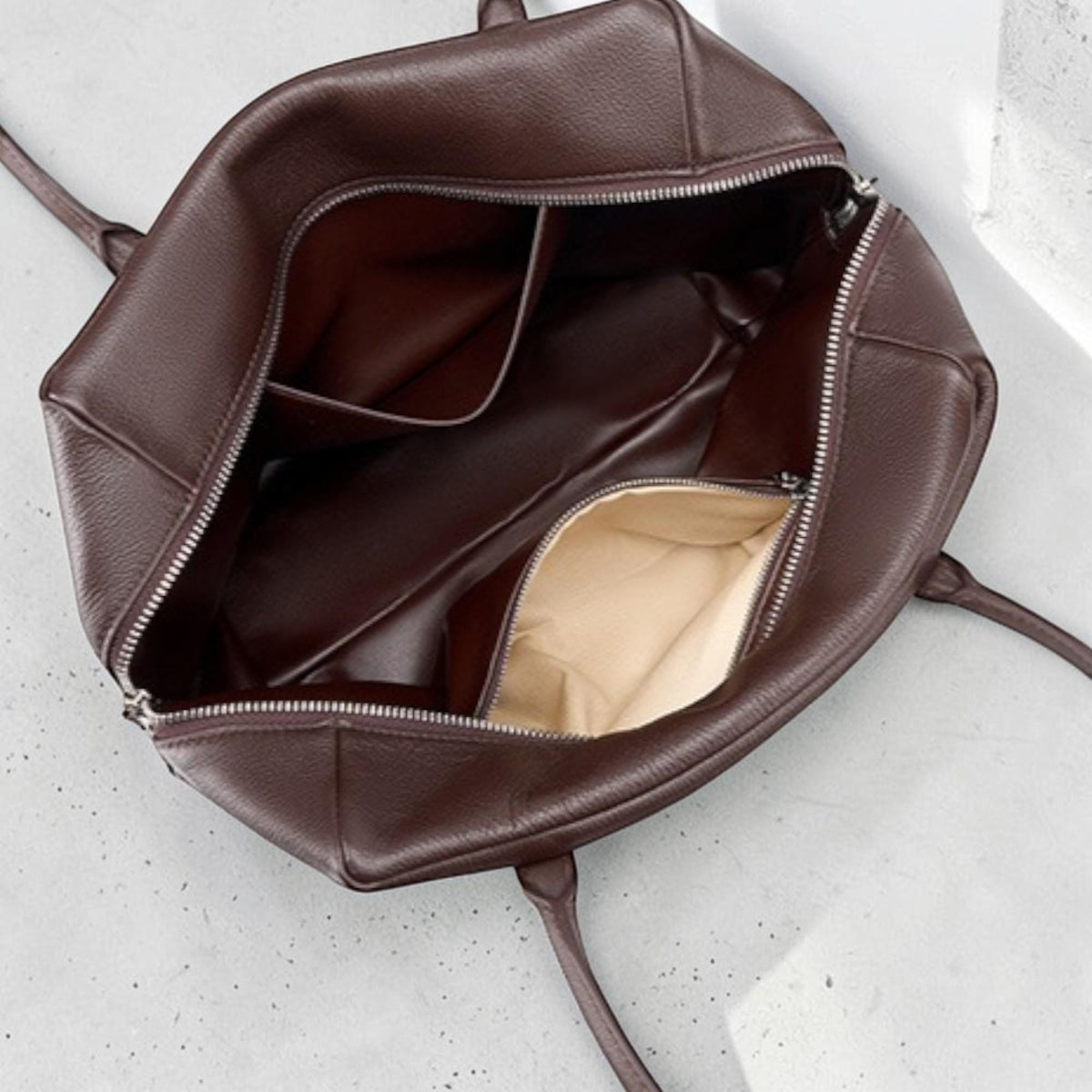 May include: Open view of a brown leather tote bag with a silver zipper. The bag has two handles and multiple interior pockets. The interior lining is a light beige color.