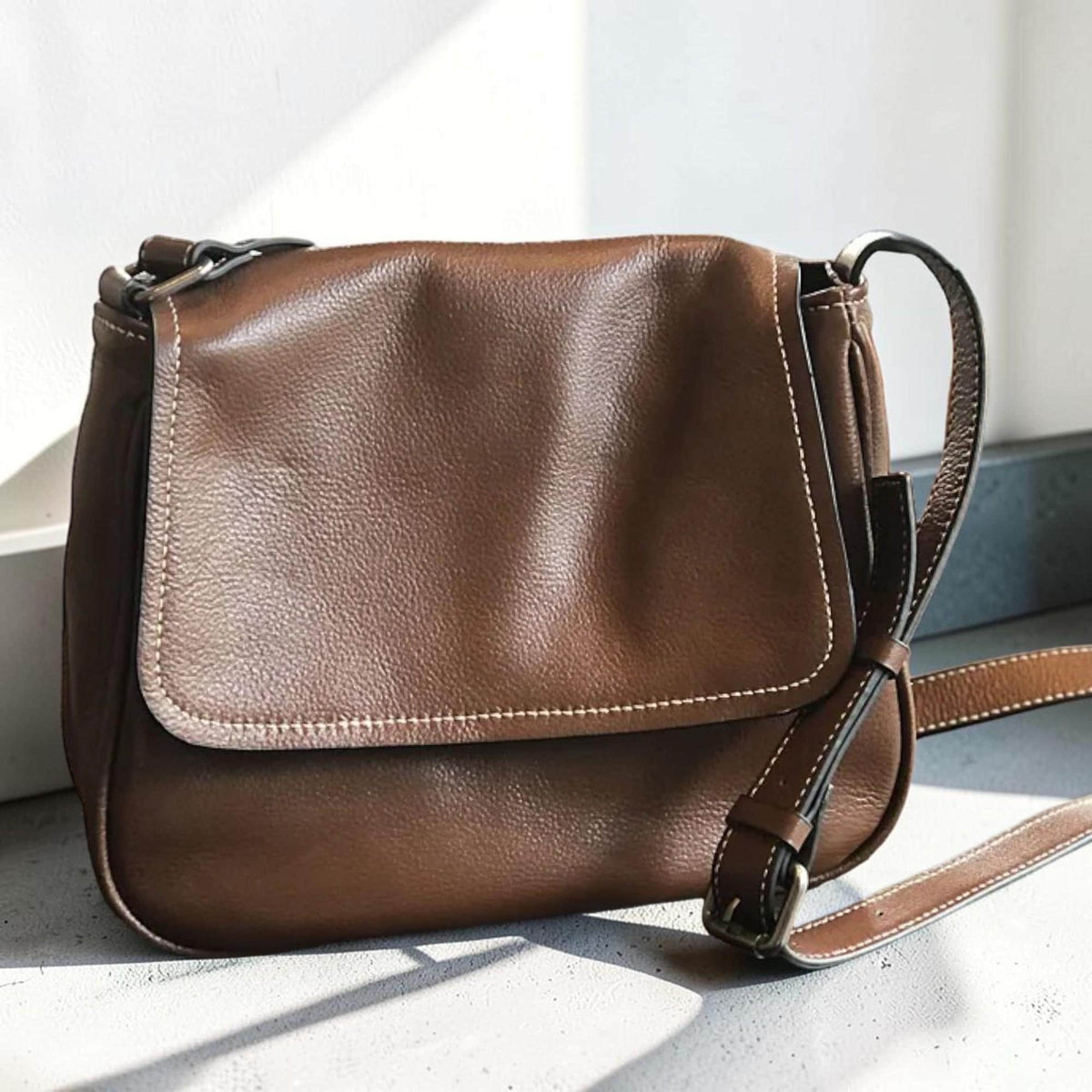 May include: A brown leather crossbody bag with a flap closure. The bag features white stitching along the edges and a long adjustable strap. The bag is made of a textured leather material.