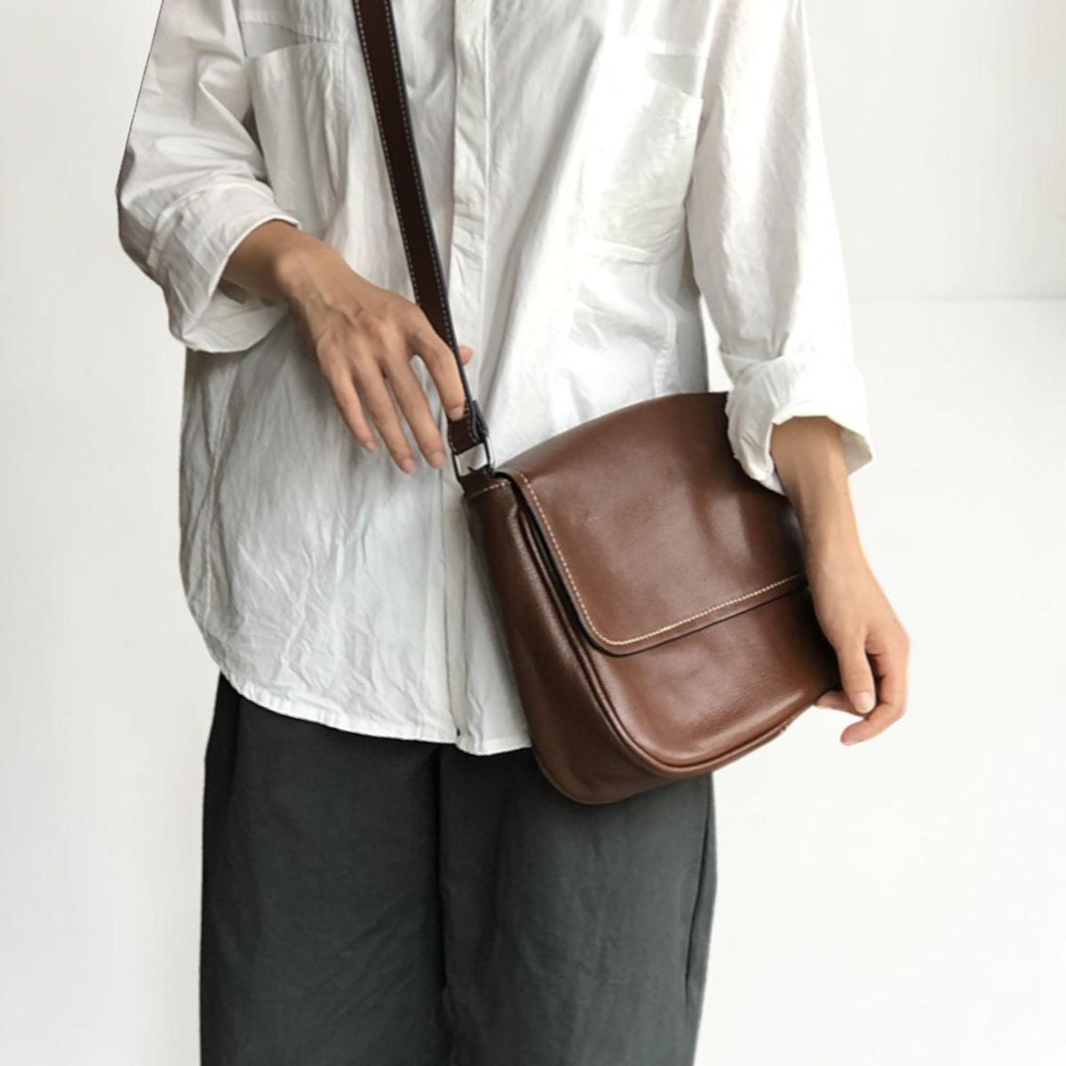 May include: A brown leather crossbody bag with a flap closure and a long adjustable strap. The bag features white stitching and a rectangular shape. The bag is worn over the shoulder.