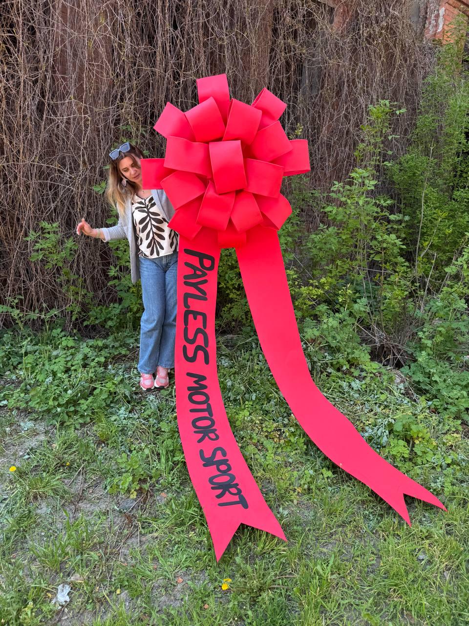May include: A large, bright red bow with long, flowing ribbons. The text "PAYLESS MOTOR SPORT" is printed in black on one of the ribbons. The bow is set against a backdrop of green foliage and grass.