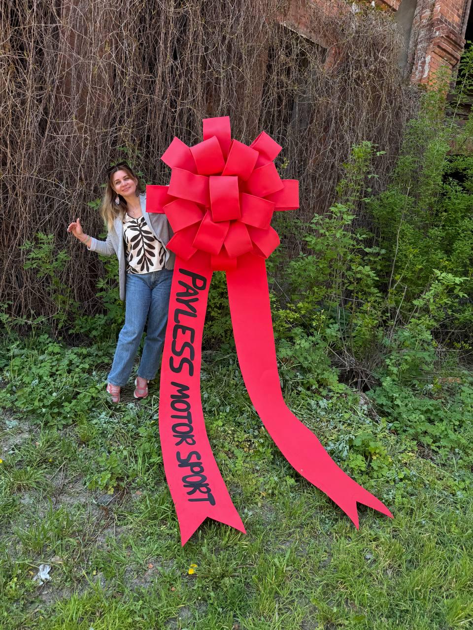 May include: A large, bright red bow with long streamers. The bow has the text "PAYLESS MOTOR SPORT" printed on one of the streamers. The bow is displayed outdoors with a person standing next to it.