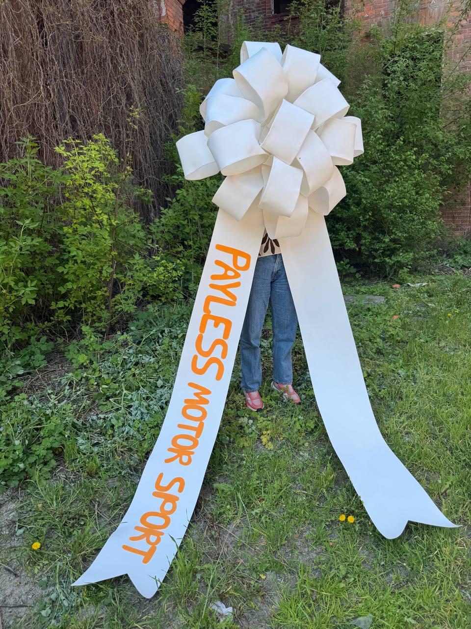 May include: A large white bow with long ribbons, featuring the orange text "PAYLESS MOTOR SPORT". The bow is made of layered paper or fabric and is displayed outdoors in front of greenery.