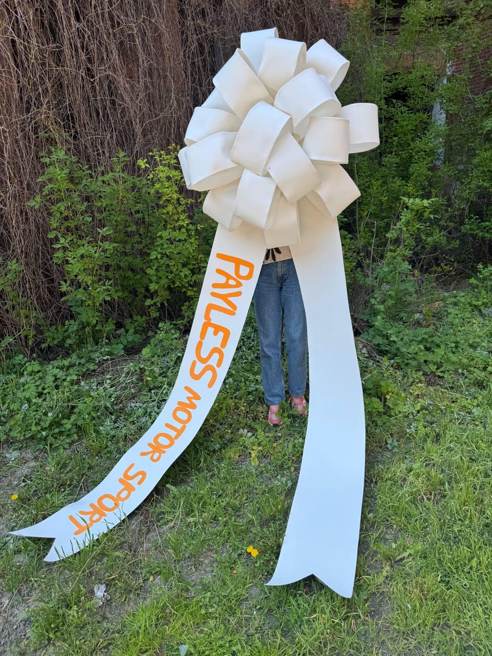 May include: A large white bow with long, flowing ribbons. The text "PAYLESS MOTOR SPORT" is printed in orange on one of the ribbons. The bow is set against a backdrop of green grass and foliage.