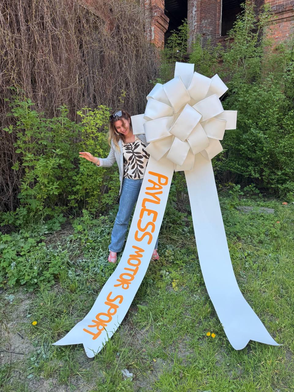 May include: A large, white decorative bow with long streamers. The text "PAYLESS MOTOR SPORT" is printed in orange on one of the streamers. The bow is set against a backdrop of greenery and an old brick building.