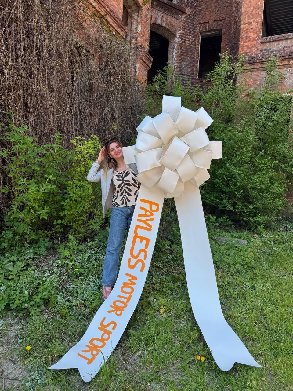 May include: A large white bow with long ribbons, featuring the text "PAYLESS MOTOR SPORT" in orange. The bow is in front of a woman wearing a patterned top and jeans, standing in front of a brick building.
