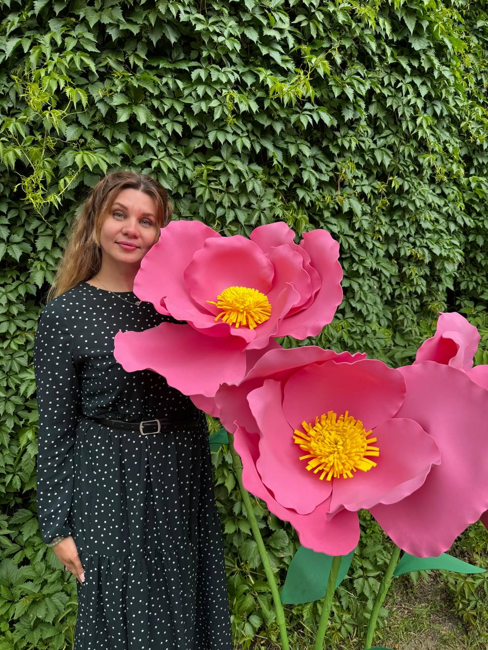 May include: Two oversized artificial pink flowers with bright yellow centers, featuring large, smooth, rounded petals. The flowers are mounted on tall green stems with green leaves, standing next to a person wearing a black dress with white polka dots.